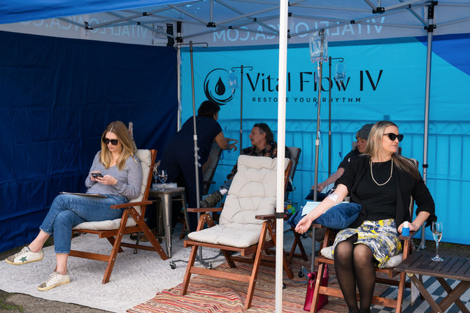 Outdoor wellness pop-up: people reclining in cushioned wooden chairs under a blue IV therapy tent receiving IV drips, a nurse attending, infusion bags and small tables with drinks in a relaxed street-side setup.