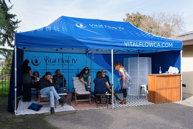 Blue branded canopy pop-up IV therapy lounge outdoors, people seated on cushioned chairs with IV poles while staff assist, small wooden bar at right and trees in the background