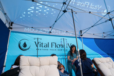 Outdoor wellness tent at an event with medical staff setting up IV therapy drips while clients relax in cushioned lounge chairs under a bright blue canopy