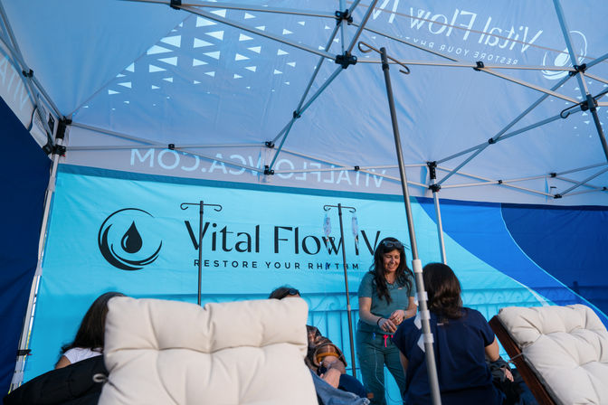 Outdoor wellness tent at an event with medical staff setting up IV therapy drips while clients relax in cushioned lounge chairs under a bright blue canopy