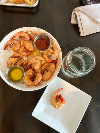 Bowl of seasoned shrimp with cocktail sauce and melted butter cups on a dark wood table, single shrimp on a square plate and a mason jar glass — ready to dip.