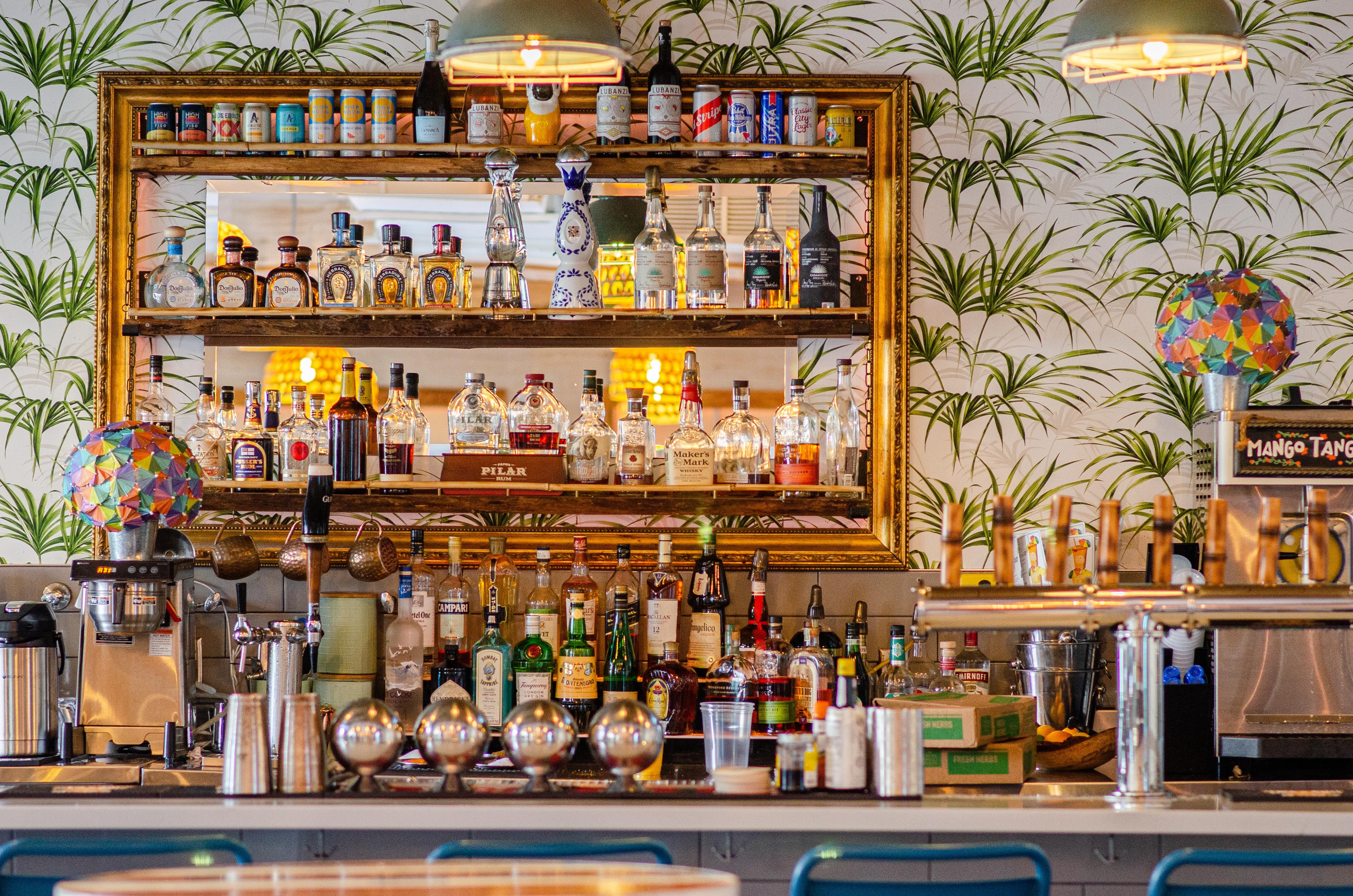 Bright modern cocktail bar interior with mirrored liquor shelves, assorted spirits and cans, palm-print wallpaper, colorful mosaic pendant lamps, and chrome draft taps on the counter.