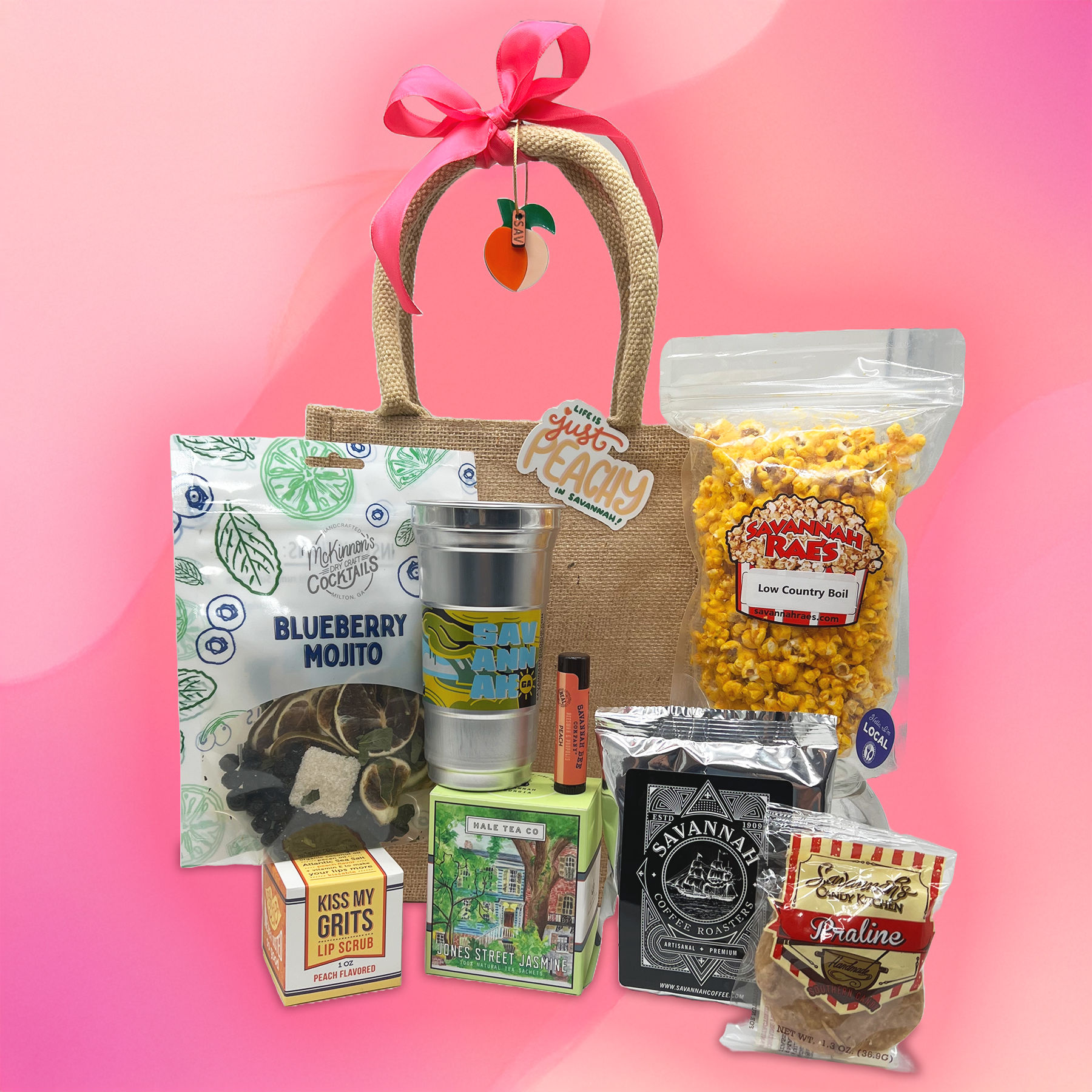 Peach-themed Georgia gift bag with pink bow showcasing a metal tumbler, bag of seasoned popcorn, blueberry mojito cocktail mix, jasmine tea box, coffee bag, peach lip scrub and praline candy on a bright pink background.