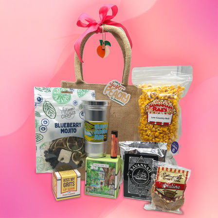 Peach-themed Georgia gift bag with pink bow showcasing a metal tumbler, bag of seasoned popcorn, blueberry mojito cocktail mix, jasmine tea box, coffee bag, peach lip scrub and praline candy on a bright pink background.