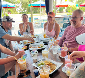 Group of friends at an outdoor patio under colorful umbrellas sharing chips, dips, and tropical cocktails with paper umbrellas, casual summer gathering