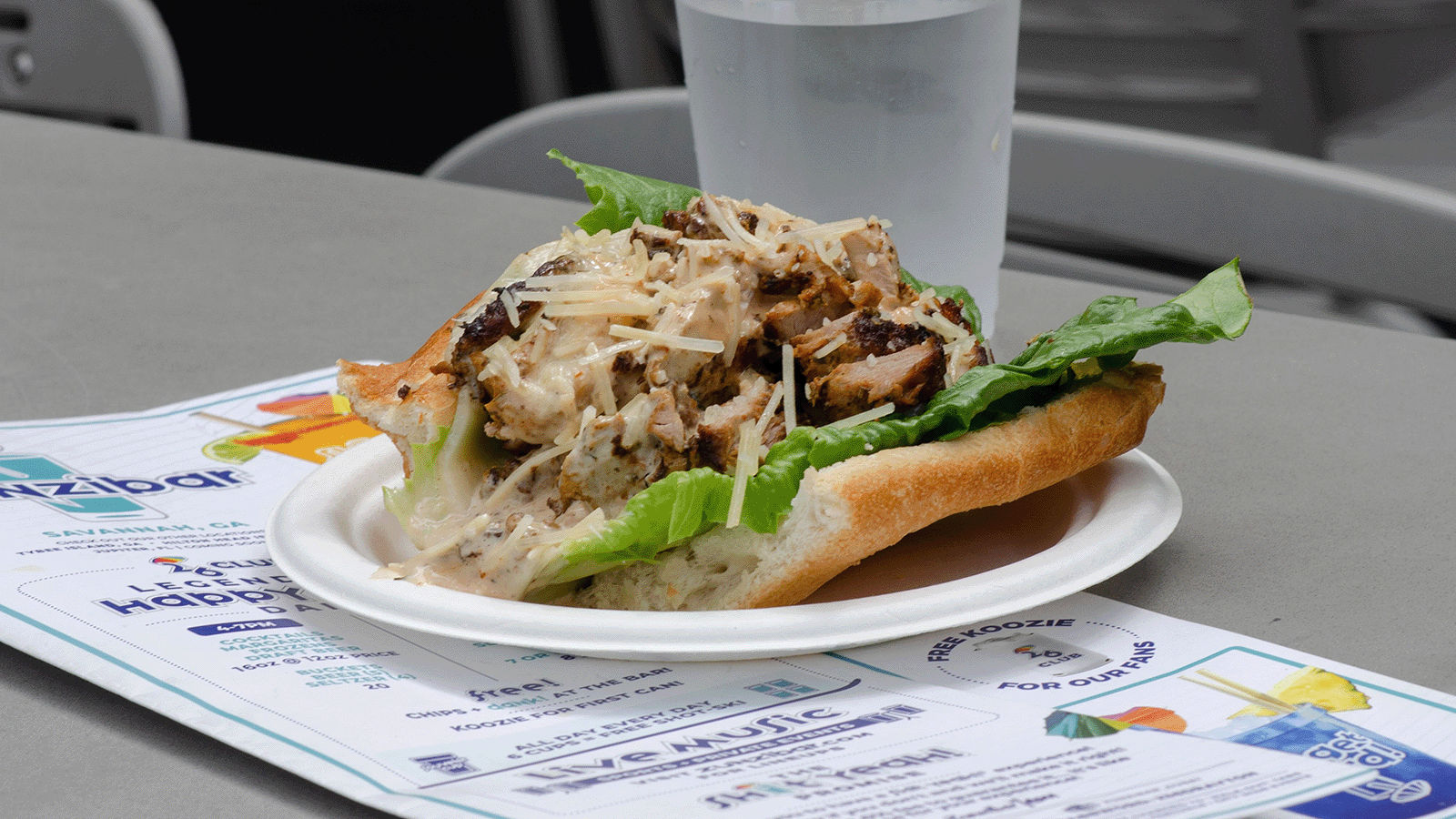 Mouthwatering grilled chicken Caesar hoagie with romaine and shaved Parmesan on a paper plate at an outdoor table, next to a water cup and menu flyer.
