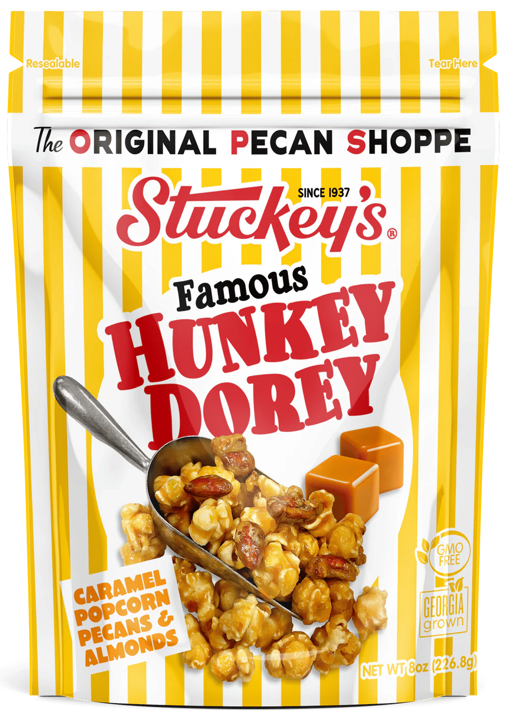 Crunchy caramel popcorn with pecans and almonds spilling from a metal scoop on a yellow-and-white striped resealable pouch, with caramel squares pictured