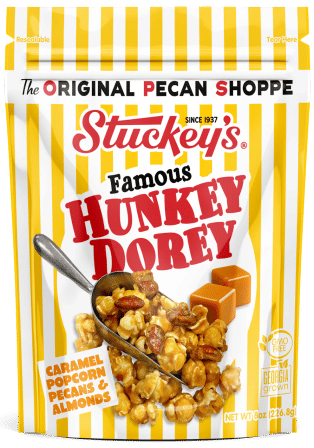 Crunchy caramel popcorn with pecans and almonds spilling from a metal scoop on a yellow-and-white striped resealable pouch, with caramel squares pictured