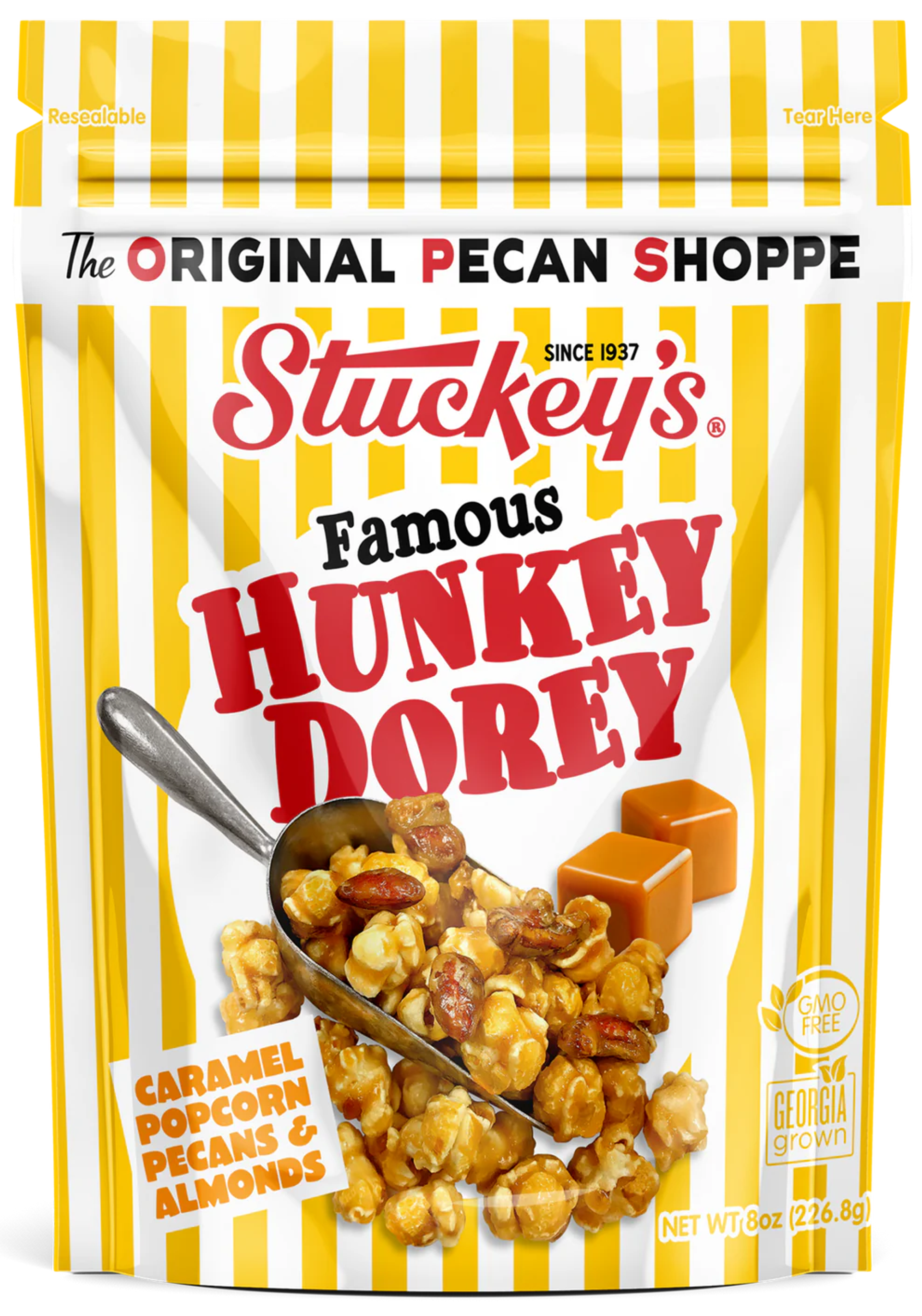 Crunchy caramel popcorn with pecans and almonds spilling from a metal scoop on a yellow-and-white striped resealable pouch, with caramel squares pictured