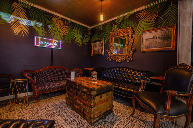 Cozy speakeasy-style lounge interior with tufted black leather sofa, patterned vintage settee, palm-leaf wall mural, ornate gilded mirror, rustic trunk coffee table, leather armchairs, warm pendant light and neon sign.