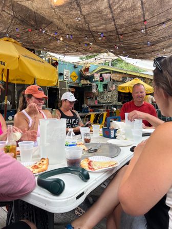 Friends enjoying pizza at a casual outdoor patio restaurant with yellow umbrellas, hanging string lights, shade canopy and eclectic surf‑shack decor and signs.