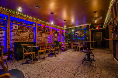Cozy lounge interior with exposed brick, purple-blue LED uplighting, string lights, tufted leather banquettes, wooden bistro chairs and a neon sign framed by greenery.