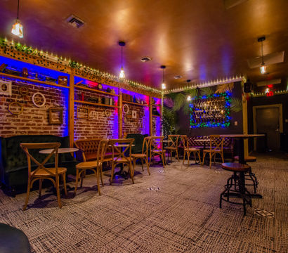 Cozy lounge interior with exposed brick, purple-blue LED uplighting, string lights, tufted leather banquettes, wooden bistro chairs and a neon sign framed by greenery.