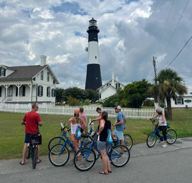 Group of people on blue beach-cruiser bikes paused by a white picket fence in front of a tall black-and-white coastal lighthouse and keeper’s houses under a dramatic cloudy sky