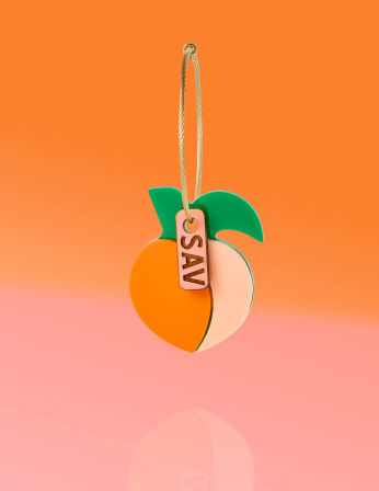 Hanging peach-shaped acrylic keychain ornament with orange and pale-pink halves, green leaves, gold cord and small metallic tag, floating on an orange-to-pink gradient background.