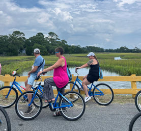 Three adults on blue cruiser bikes paused on a yellow wooden bridge over a coastal salt marsh with a cloudy sky and tree-lined horizon