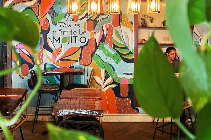 Cozy plant-framed cafe interior with a colorful mural reading "This is mint to be MOJITO", warm pendant lights, rustic wooden tables and a seated customer.