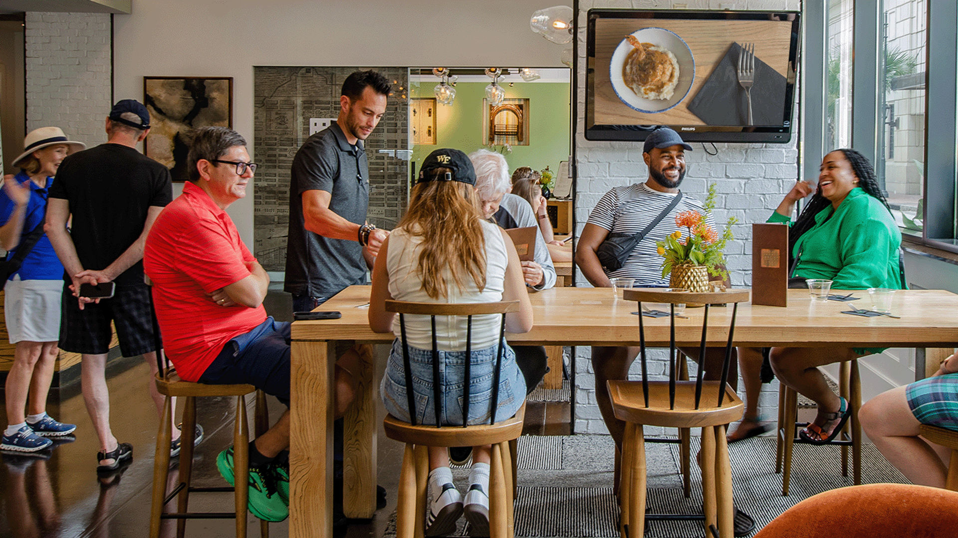 Bright urban cafe interior with a group seated at a long wooden communal table, chatting and laughing as a server takes orders; TV on the wall and a small floral centerpiece add cozy vibe.