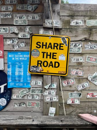 Rustic wooden wall covered with signed U.S. dollar bills, a bright yellow “SHARE THE ROAD” sign, stickers and a blue drink poster — playful roadside bar décor.