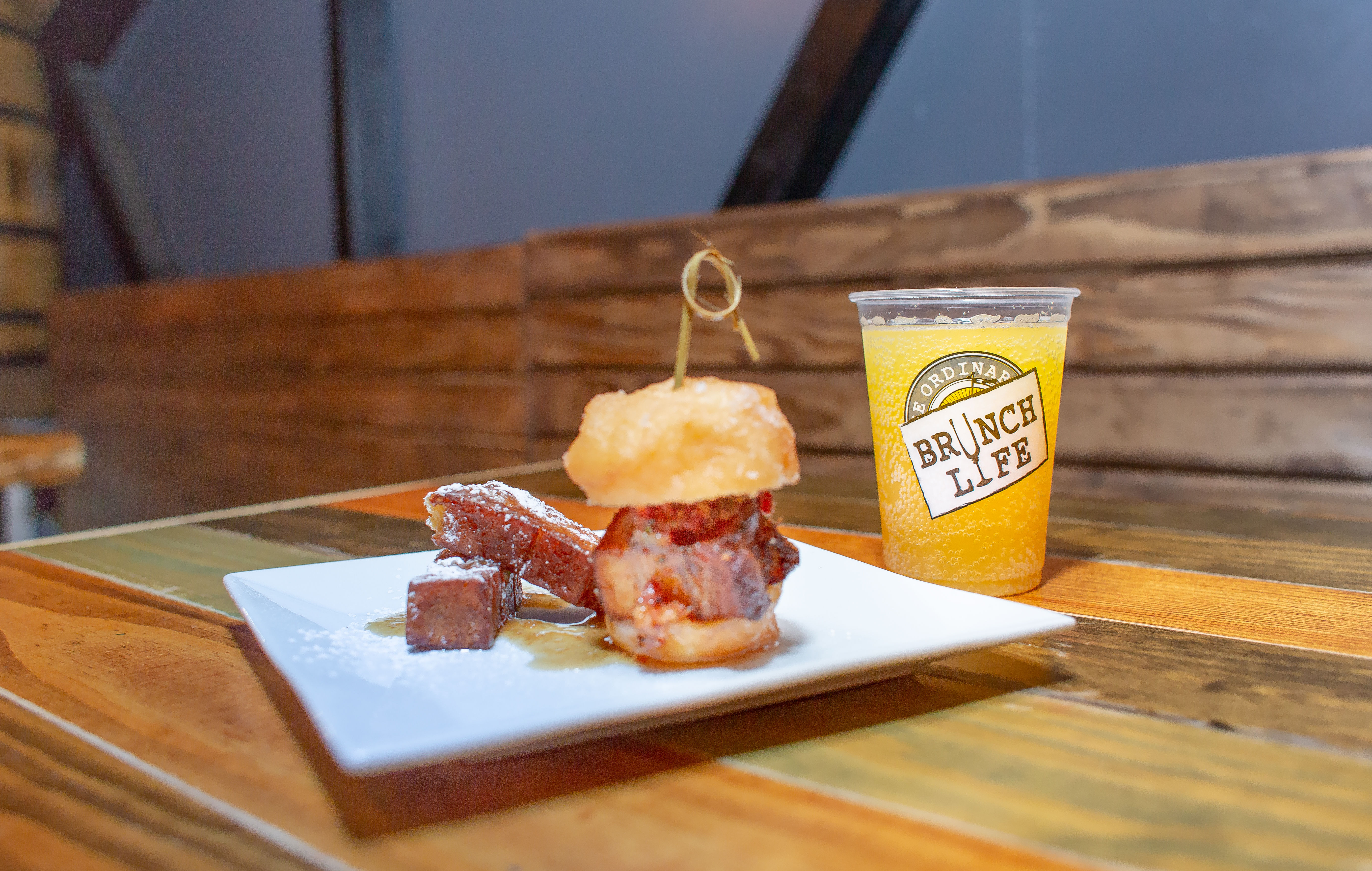 Brunch plate with glazed pork belly cubes and a skewered mini-donut slider on a white plate, beside an orange brunch drink in a plastic cup on a wooden table