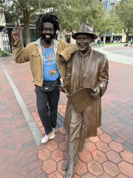 Smiling person with dreadlocks in a tan jacket and blue tour shirt flashing a peace sign while posing with a bronze statue of a suited man holding a newspaper on a brick city plaza with trees and buildings in the background