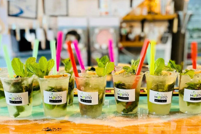 Row of mojito cocktails in clear plastic cups on a bar counter, garnished with fresh mint leaves, lime wedges, crushed ice and bright colorful straws for a casual tropical bar vibe.