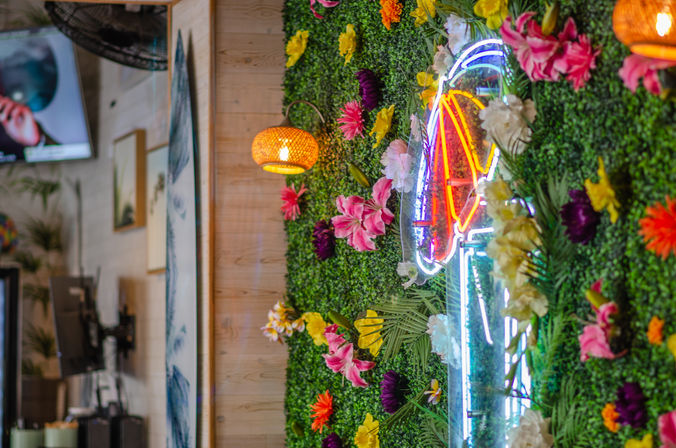 Vibrant tropical-themed cafe interior with a green faux foliage wall studded with colorful flowers, a neon umbrella sign, rattan pendant lights and warm wooden accents.
