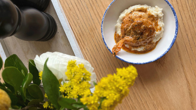 Blue-rimmed bowl of creamy mashed potatoes topped with a saucy shrimp on a wooden table, framed by bright yellow flowers and a black pepper mill.