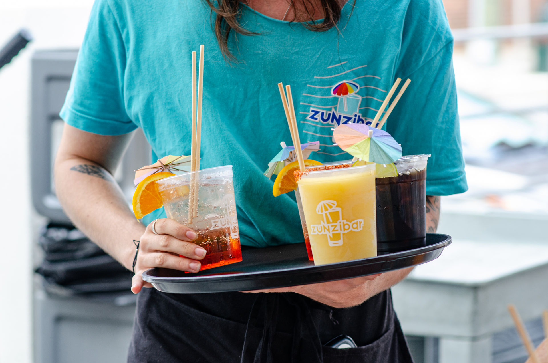 Server holding a tray of colorful tropical drinks in clear plastic cups with paper umbrellas, orange slices and straws on an outdoor patio, wearing a teal shirt