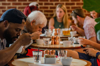 Busy brick-walled café with patrons at round tables enjoying soup from white bowls, glasses of water and copper salt-and-pepper shakers on the table.