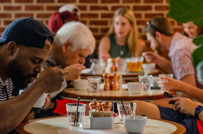 Busy brick-walled café with patrons at round tables enjoying soup from white bowls, glasses of water and copper salt-and-pepper shakers on the table.