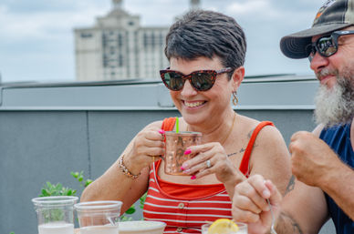 Smiling woman in a red striped tank top and sunglasses sips from a copper mug with a green straw on a sunny rooftop patio, laughing with a bearded friend as drinks and plates sit on the table and a city building blurs in the background.