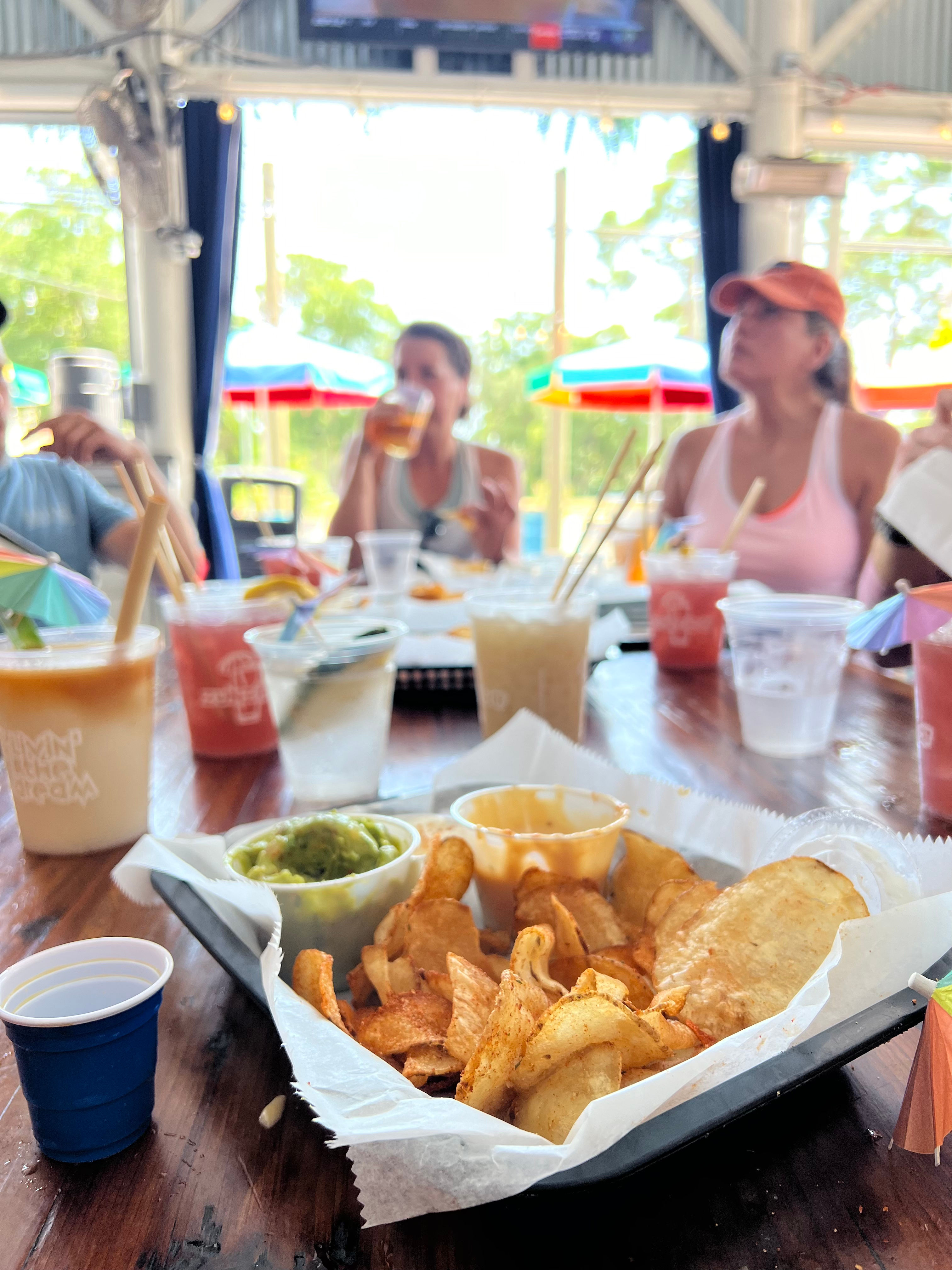 Sunny outdoor patio scene: basket of crispy potato chips with guacamole and dipping sauce on a wooden table, surrounded by colorful cocktails and people enjoying drinks