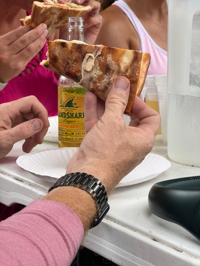 Close-up of hands holding a cheesy, charred-crust pizza slice over a paper plate next to a yellow-labeled beer bottle, plastic cups and a condensation-covered water pitcher on a white folding table at a casual summer gathering.