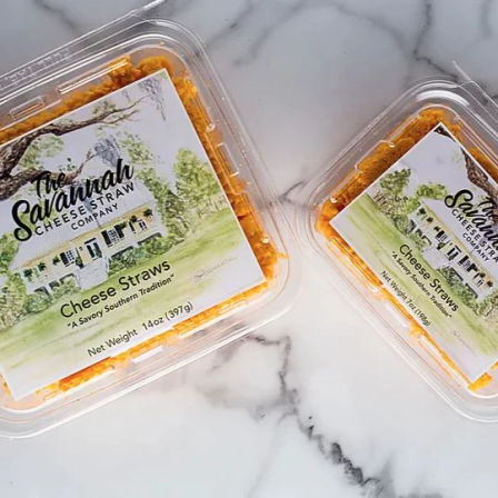 Two clear plastic containers of orange Southern-style cheese straws with illustrated labels on a white marble countertop.