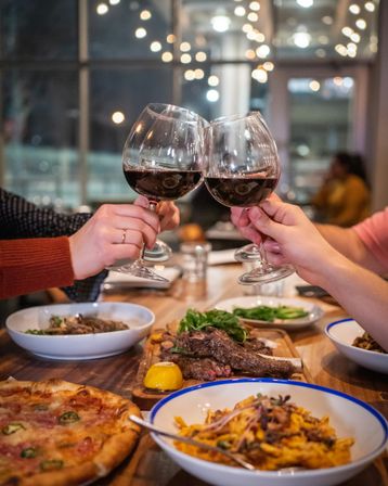 Hands clinking red wine glasses over a wooden table with steak, pizza and pasta in a cozy restaurant under string lights