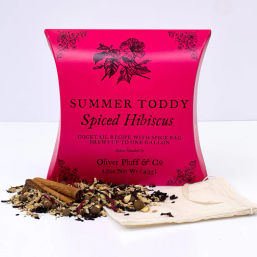 Hot-pink pillow box of spiced hibiscus toddy cocktail mix with loose dried hibiscus and whole spices, cinnamon sticks, and a reusable muslin spice bag on a white background.