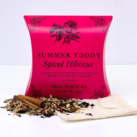 Hot-pink pillow box of spiced hibiscus toddy cocktail mix with loose dried hibiscus and whole spices, cinnamon sticks, and a reusable muslin spice bag on a white background.