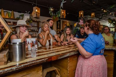 Group of customers sampling drinks at a rustic wooden tasting counter inside a tropical‑themed shop with hanging lanterns and product shelves.