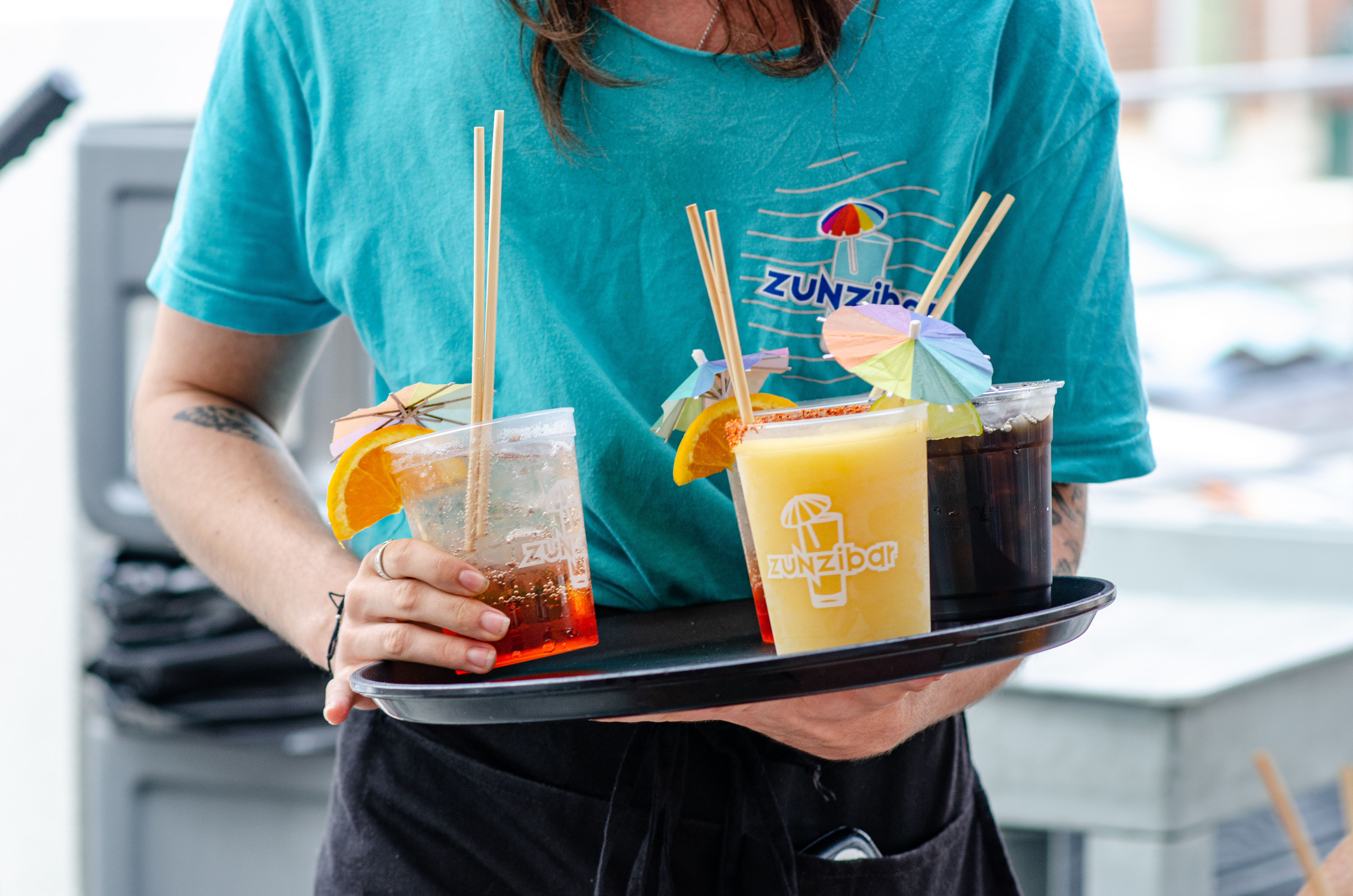 Server holding a tray of colorful summer drinks—frozen mango slush, dark cola, and a sparkling cocktail—garnished with orange slices and paper umbrellas, wearing a teal shirt with a beach-themed logo on an outdoor patio.