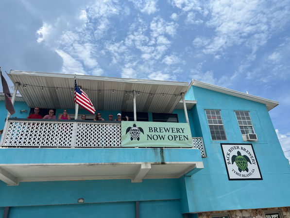 Cheerful turquoise two-story building with a second-floor balcony of people, an American flag, and a large 'Brewery Now Open' sign under a bright blue sky with scattered clouds.