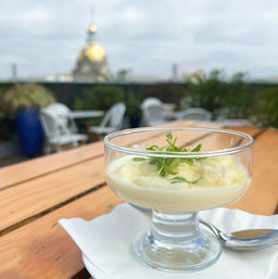 Lemon mousse in a glass coupe topped with microgreens on a wooden rooftop table, blurred terrace seating and a gold-domed city building in the background.