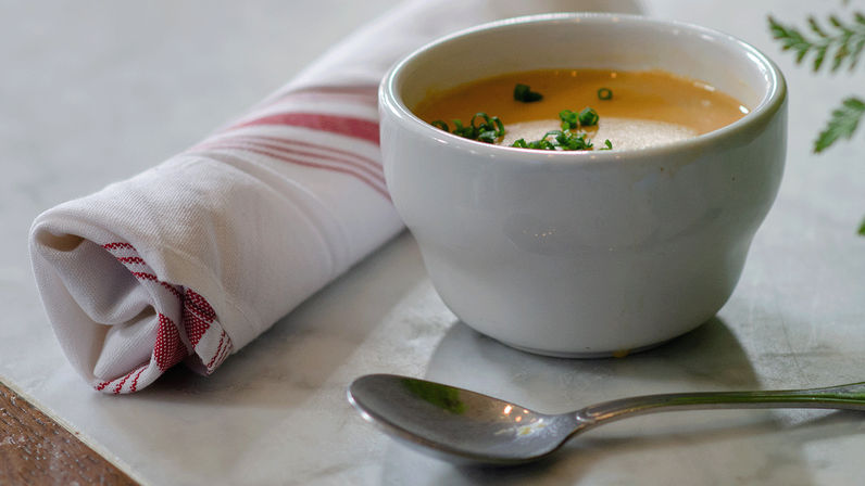 Creamy orange soup garnished with chopped chives in a white bowl on a marble table, beside a rolled red-striped napkin and a spoon — cozy bistro table setting