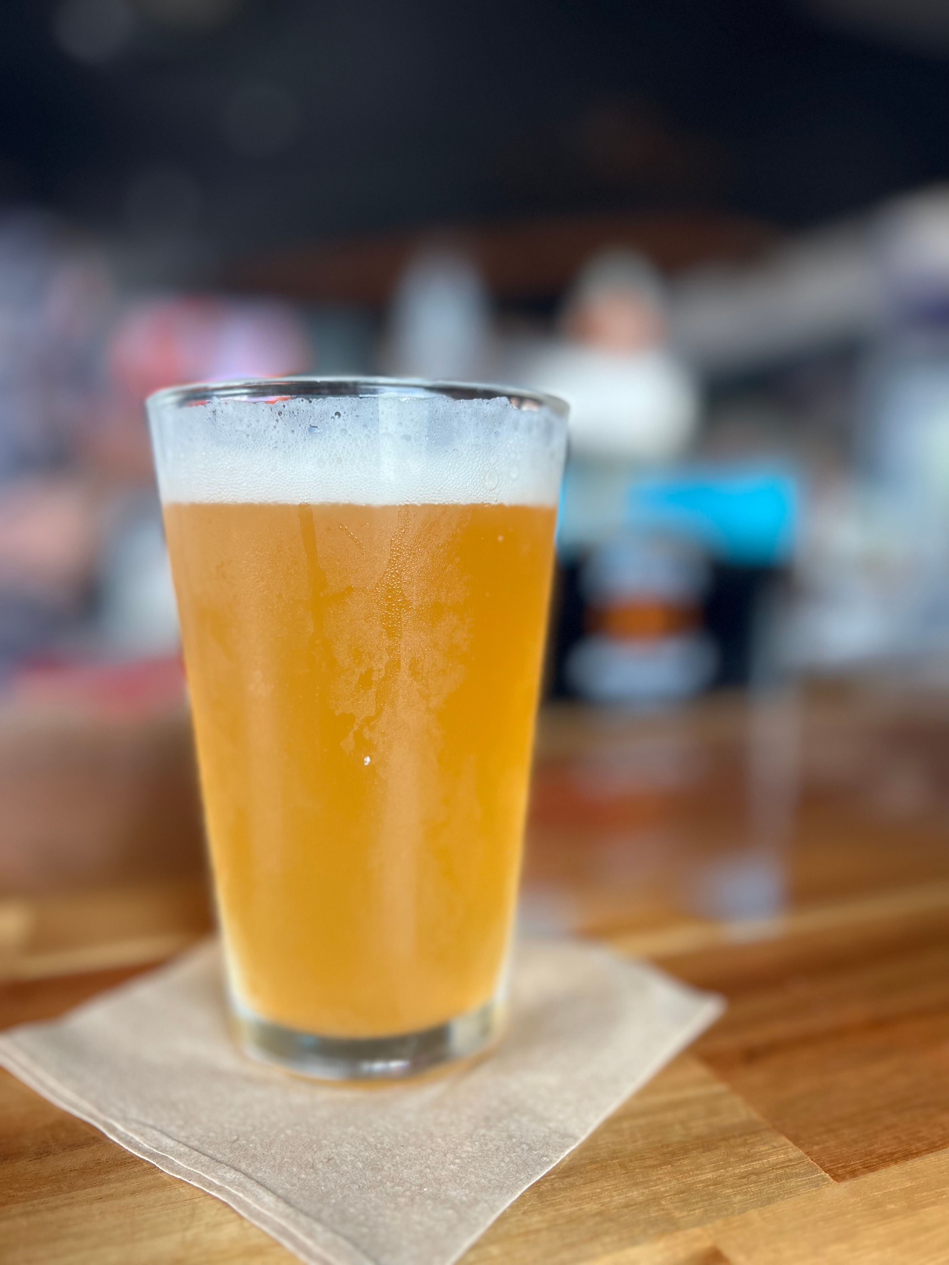 Close-up of a hazy golden craft beer in a pint glass with a foamy head on a napkin atop a wooden bar, blurred taproom background.