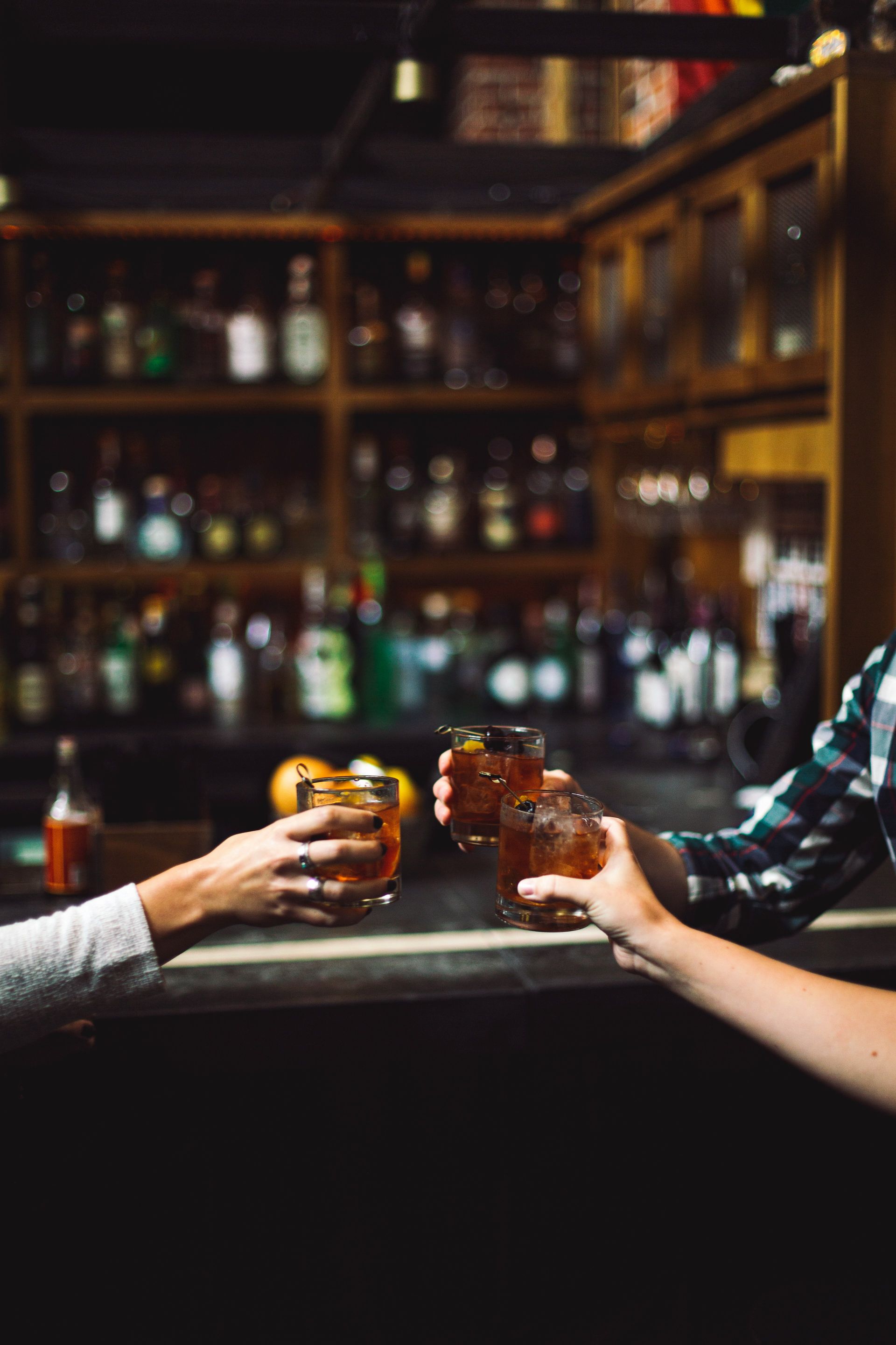 Three hands clinking whiskey cocktails over a dim wooden bar with blurred bottles on shelves — cozy urban craft cocktail bar vibe.