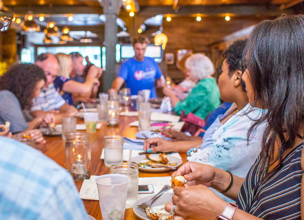 Casual group dinner at a long wooden table in a cozy indoor restaurant, people sharing plates and drinks under warm string lights — lively family-style dining scene.