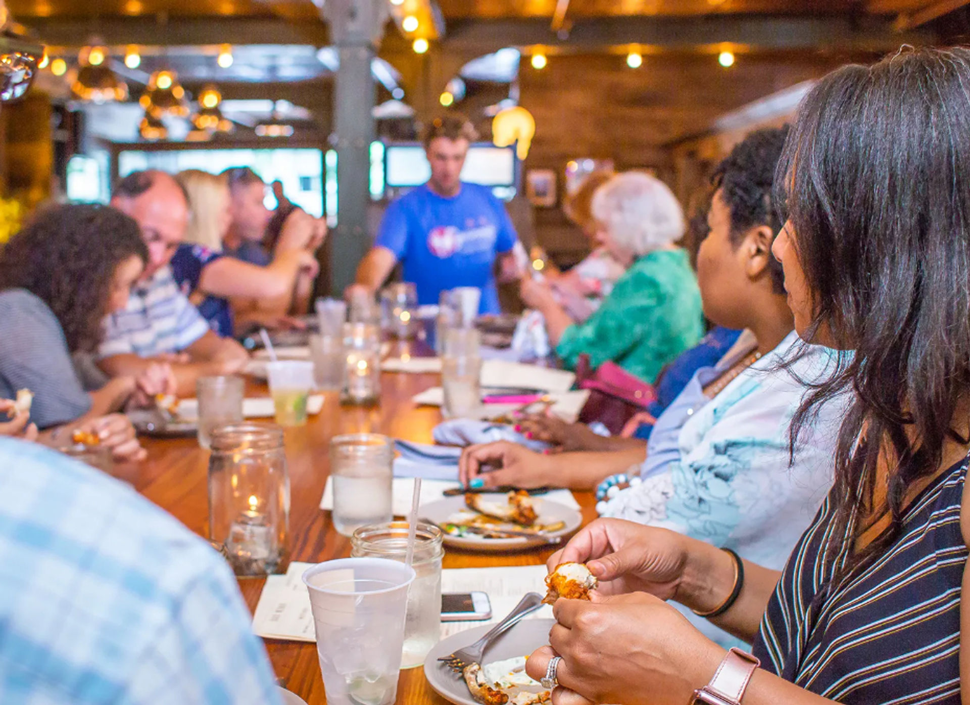 Casual group dinner at a long wooden table in a cozy indoor restaurant, people sharing plates and drinks under warm string lights — lively family-style dining scene.
