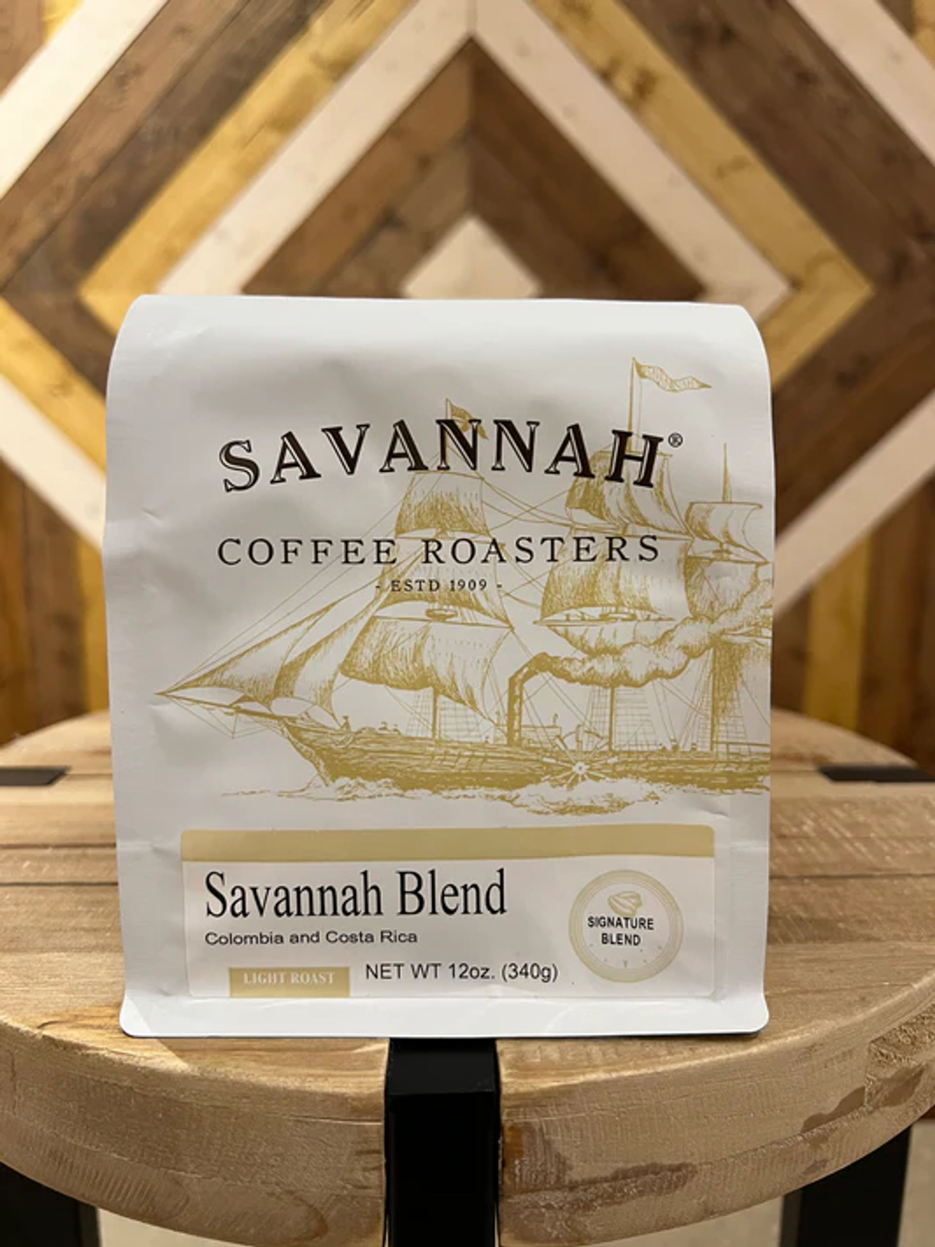 White 12 oz light-roast coffee bag from Colombia and Costa Rica displayed on a round wooden stool in front of a chevron wood-panel wall.