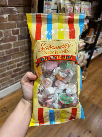 Hand holding a yellow striped bag of assorted salt water taffy with a clear window showing individually wrapped pastel candies, photographed inside a candy shop with brick wall and wooden floor.