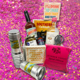 Party-ready cocktail gift set with mini bottles and mixers, jarred garnishes, stacked metal cups, playful napkins and a spiced toddy packet on a sparkly pink confetti backdrop.